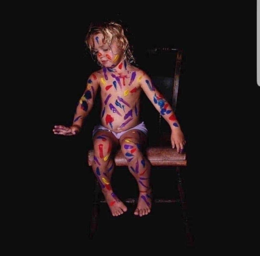 A photograph of a young child marked with various colors of paint covering nearly the entire body.